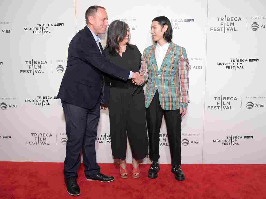 Chestnut and Kobayashi shake hands on a red carpet, with a woman standing in between them.