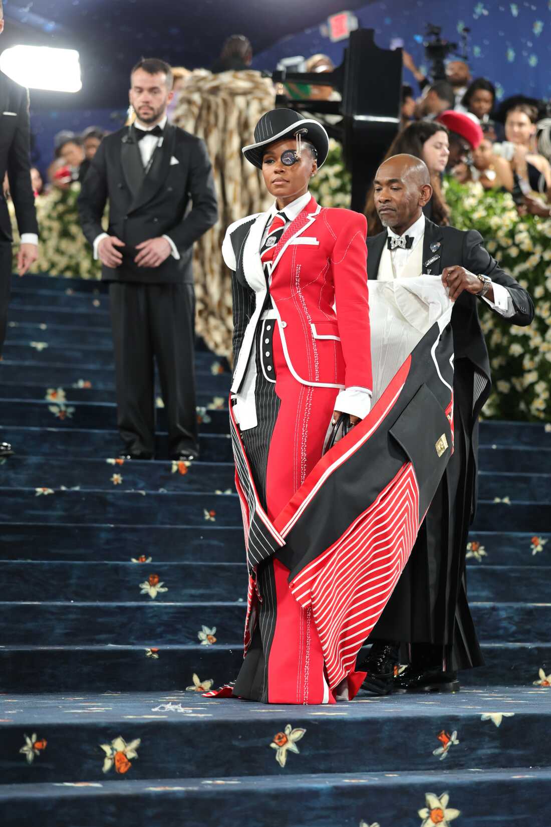 Janelle Monáe, wearing a floor-length overcoat is unzipped to reveal a suit that is half black and half red, attends the 2025 Met Gala.